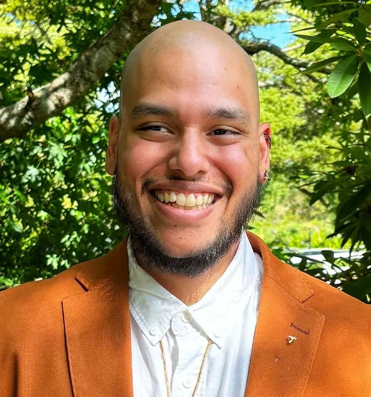 A smiling man with a shaved head and beard wearing a white shirt and an orange blazer, standing outside with green trees and leaves in the background.