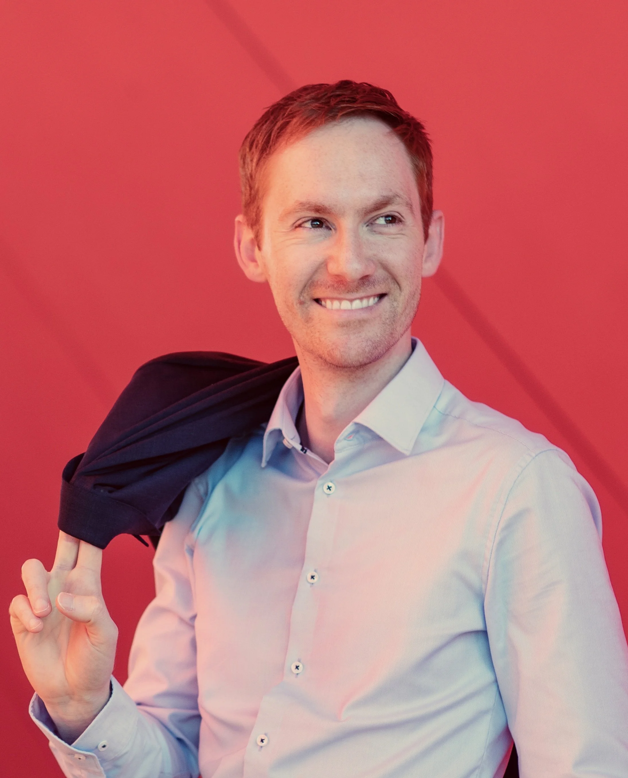 A smiling man with short red hair wearing a light-colored shirt and carrying a dark jacket over his shoulder in front of a red background.
