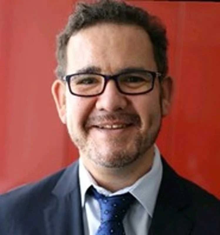 Man with short brown hair, glasses, and a beard, smiling in front of a red background, wearing a suit and tie.