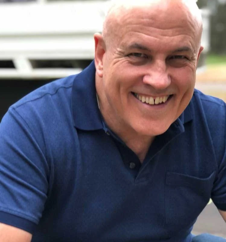 A smiling bald man with a mustache wearing a navy blue polo shirt, outdoors in front of a white vehicle.