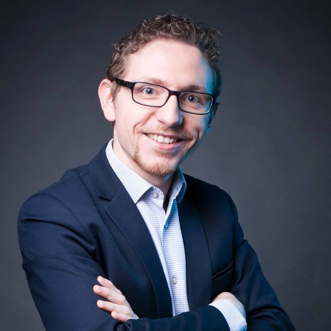 Headshot of a smiling man wearing glasses, a navy blazer, and a light collared shirt, with crossed arms against a dark background.