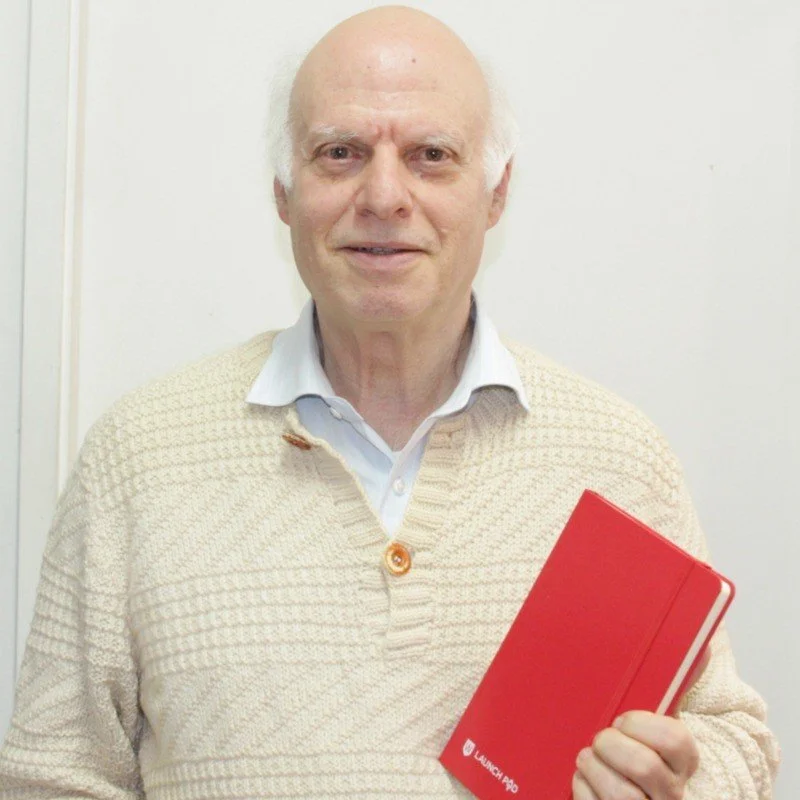 An elderly man with white hair and fair skin smiling and holding a red notebook or planner in his right hand, standing against a plain white wall.