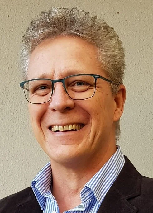 A smiling middle-aged man with gray hair, glasses, and a blue striped shirt, wearing a dark blazer, standing in front of a neutral wall.