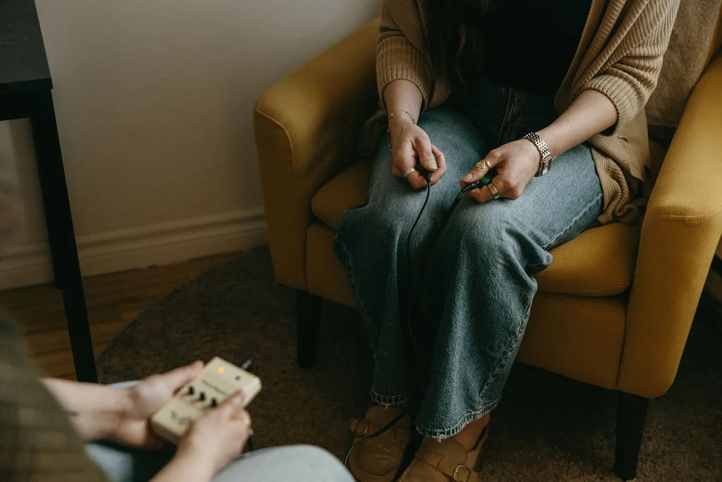 A person sitting on a yellow armchair holding a video game controller, playing a game, while another person holds a different gaming controller in front of them.
