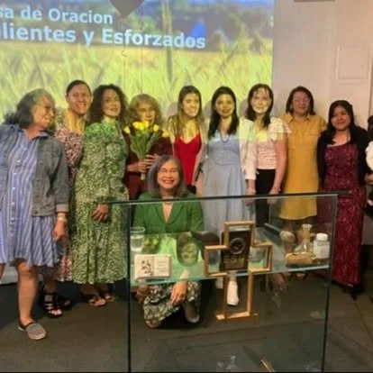Group of nine women standing together indoors, with one woman kneeling in front, holding a bouquet and smiling. Behind them is a screen with Spanish text.