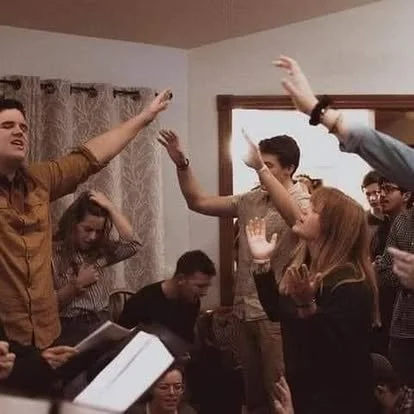 Group of young adults in a room raising their hands, some singing or praying, possibly in a communal or spiritual activity.