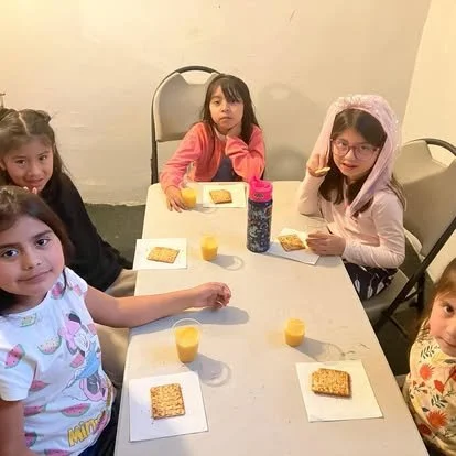 Four young girls sitting around a table with breakfast items, including juice and granola bars, in a room with plain walls.