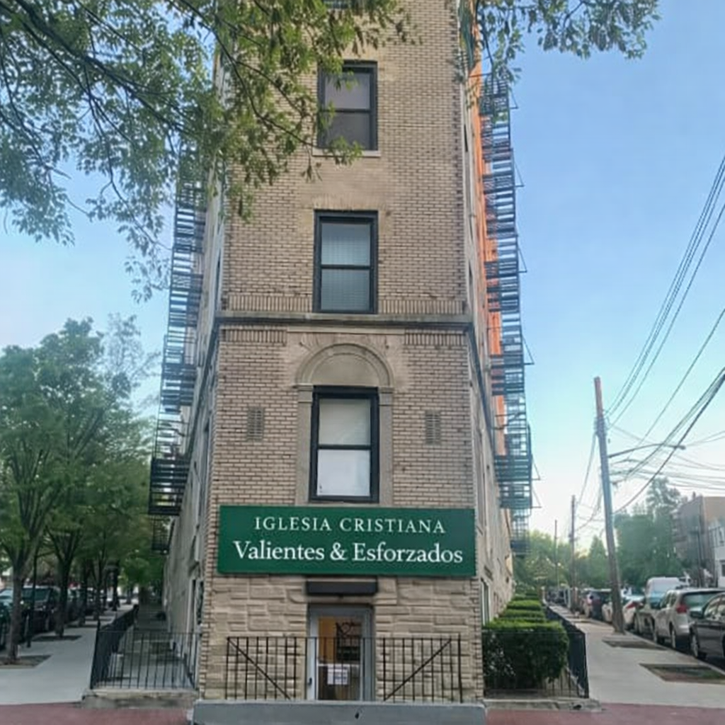 A three-story brick building with a green sign that reads 'Iglesia Cristiana Valientes & Esfuerzo' beneath a central arched decorative element. The building has black window frames and a fire escape on the right side, with trees and parked cars along the street.
