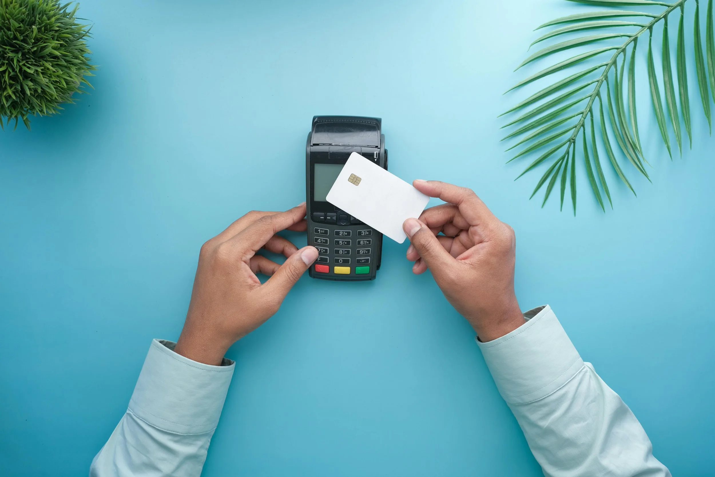 Person holding a white credit card near a black point-of-sale terminal on a light blue surface, with green plants on either side.