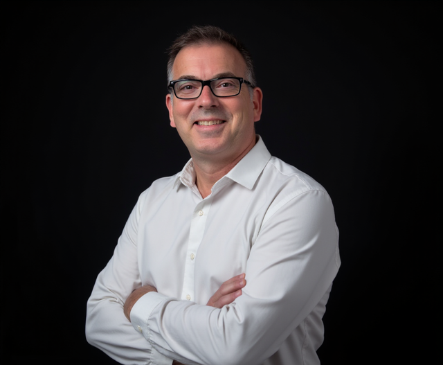 A man with glasses, wearing a white shirt, smiling with arms crossed, against a black background.