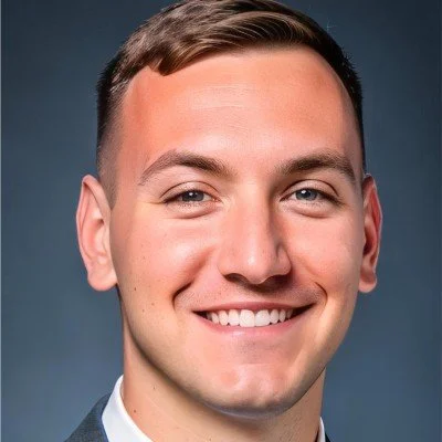 A young man with short brown hair, smiling, wearing a suit and tie, with a plain dark background.