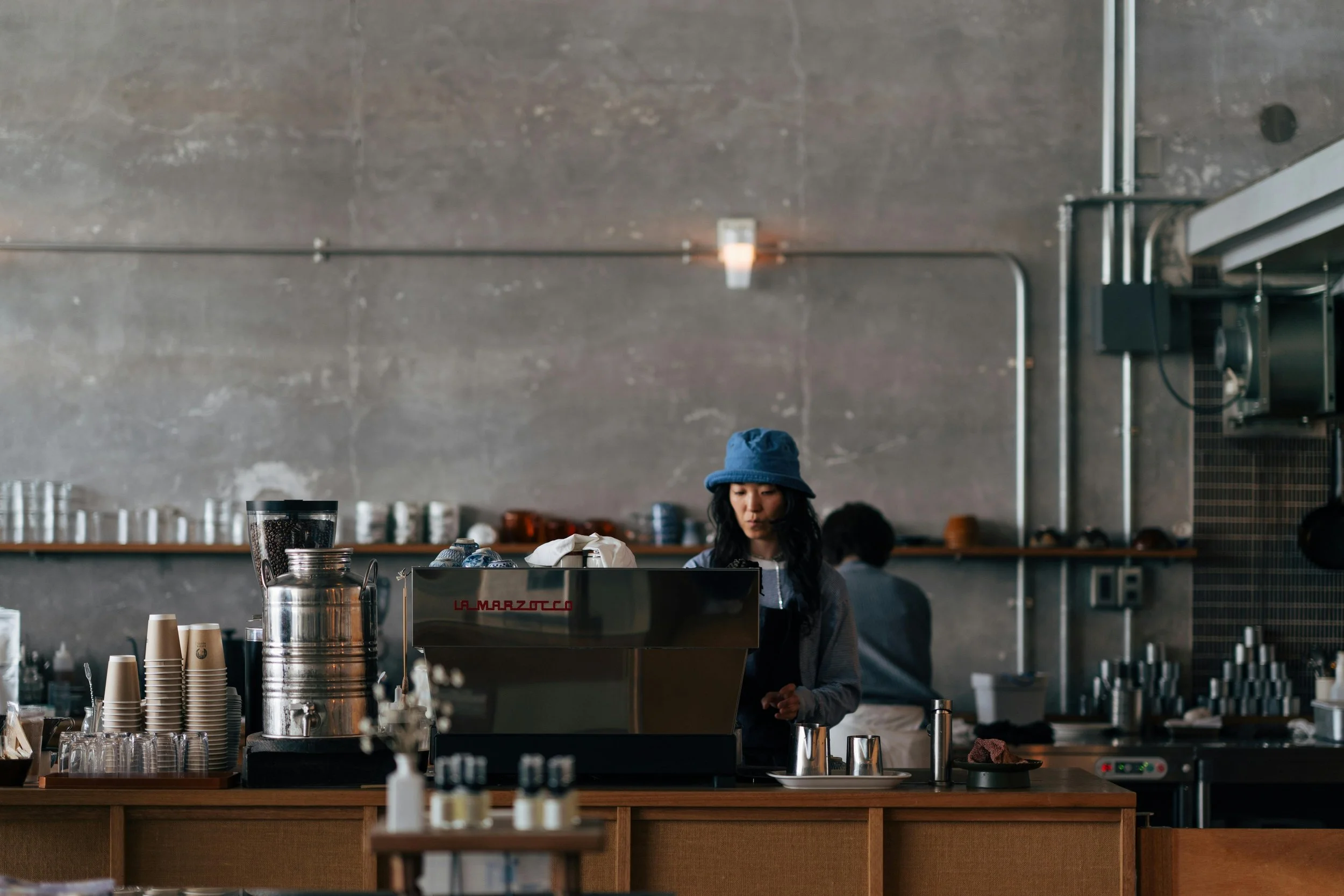 A woman with a blue bucket hat working behind a coffee counter in a cafe with a gray industrial-style wall.