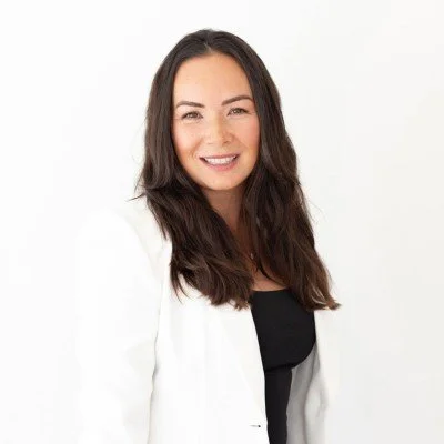 A smiling woman with long dark hair, wearing a white blazer over a black top, against a white background.