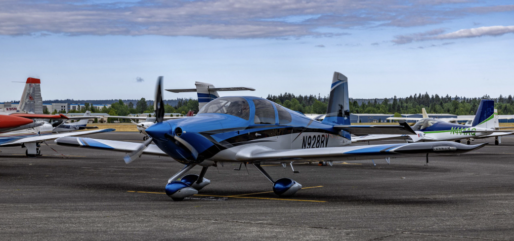 A small blue, black, and white propeller airplane parked on an airport tarmac with other small aircraft in the background and trees under a partly cloudy sky.