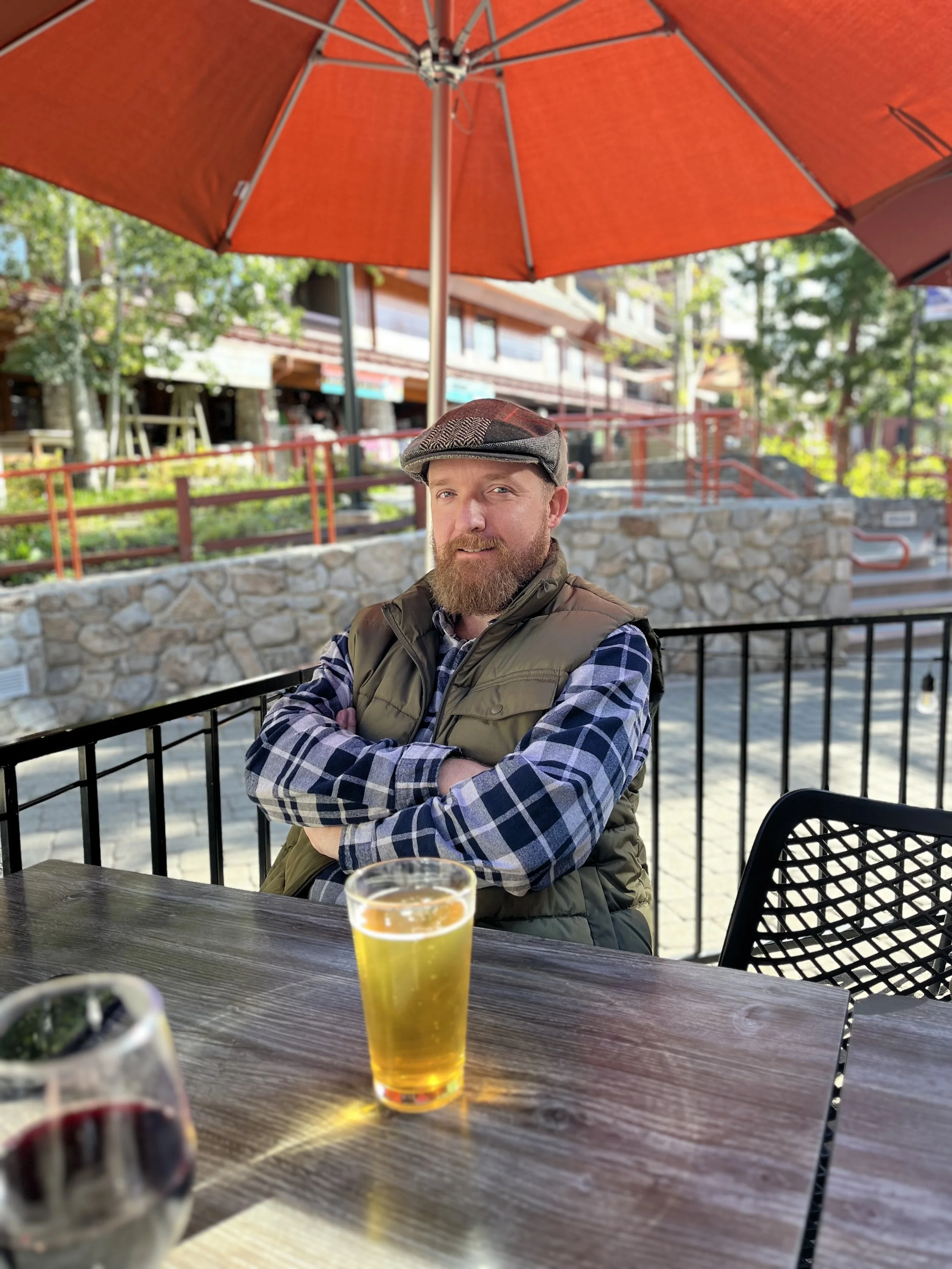 A man with a beard, wearing a flat cap and a plaid shirt with a vest, sits at an outdoor table with a glass of beer and a glass of red wine. He is under a large orange patio umbrella, in front of a stone wall and a fence, with trees and buildings in the background.