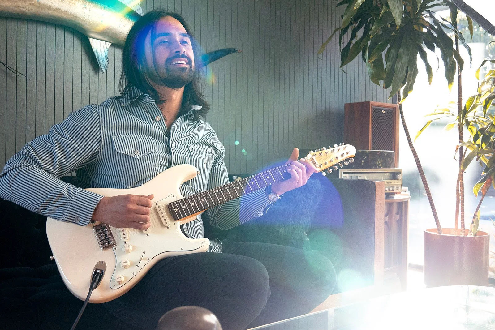 Man with long hair and beard playing an electric guitar indoors, sunlight streaming in, plants and vintage audio equipment in the background.