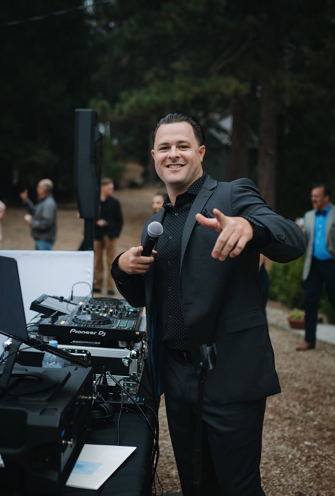 A man dressed in black suit holding a microphone and pointing towards the camera at an outdoor event, with a DJ setup on a table in front of him and several people in the background.