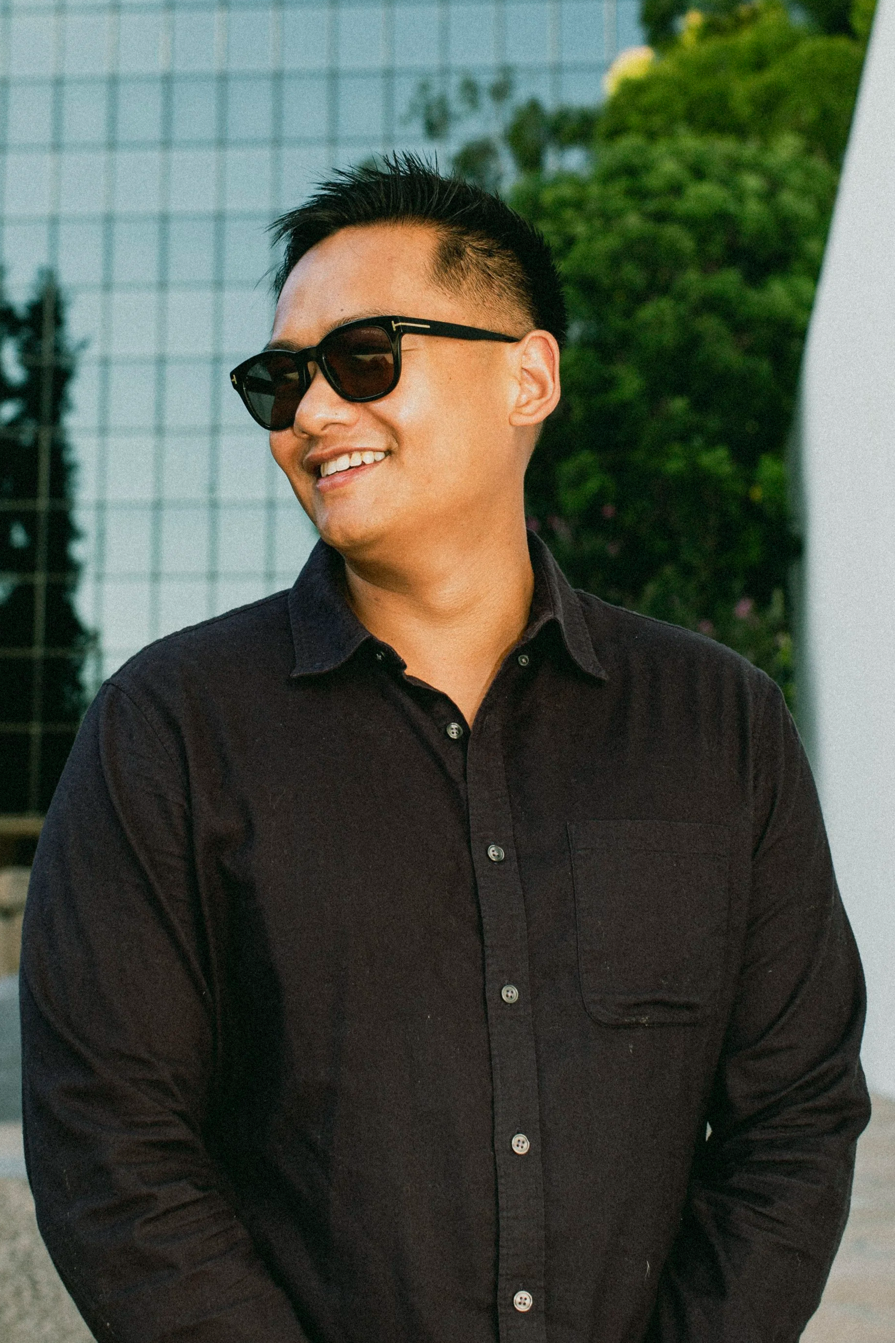 Young man wearing black sunglasses and a black button-up shirt, smiling outdoors with a modern glass building and greenery in the background.