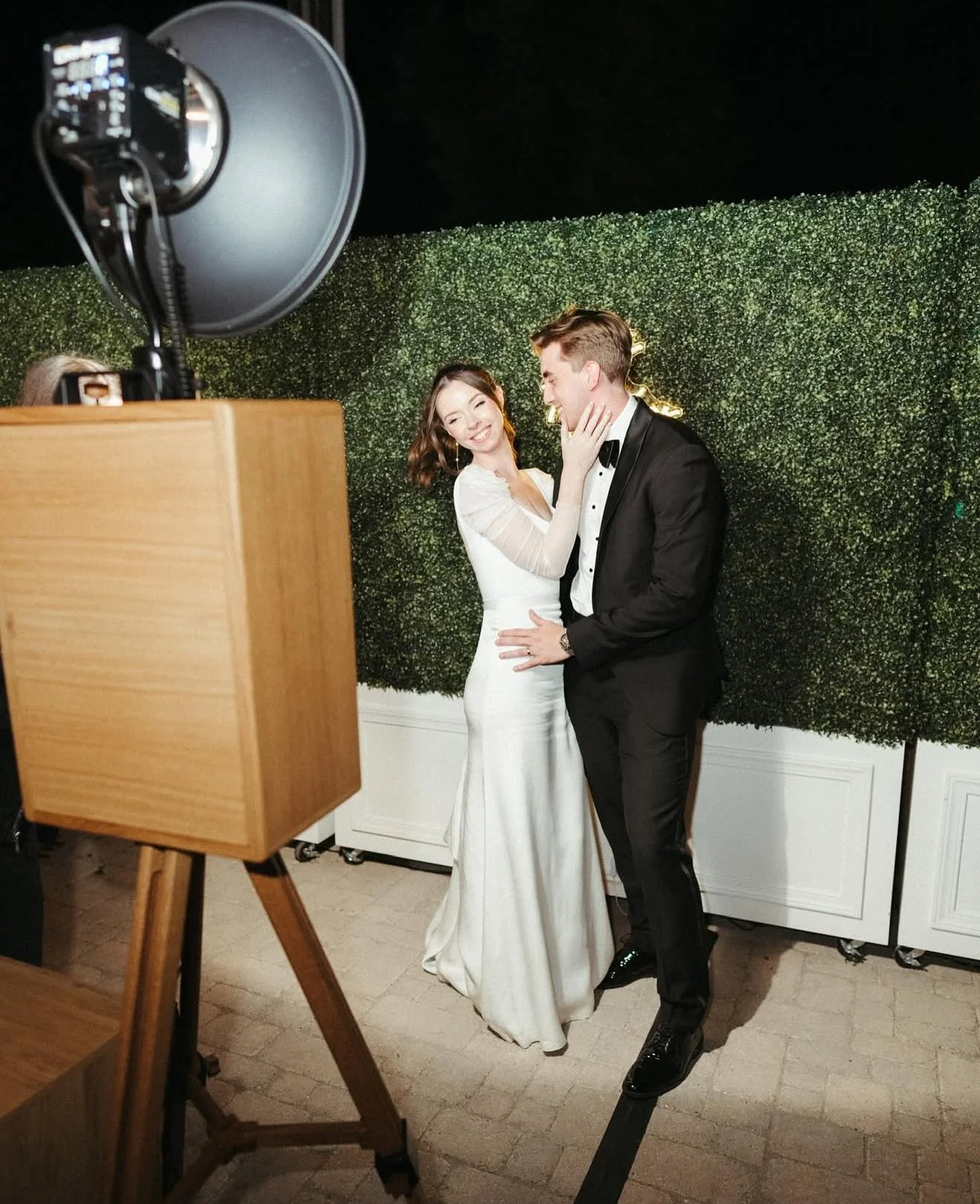 A bride and groom sharing a joyful moment at their wedding photoshoot, standing in front of a greenery backdrop, with a camera on a tripod capturing the scene.