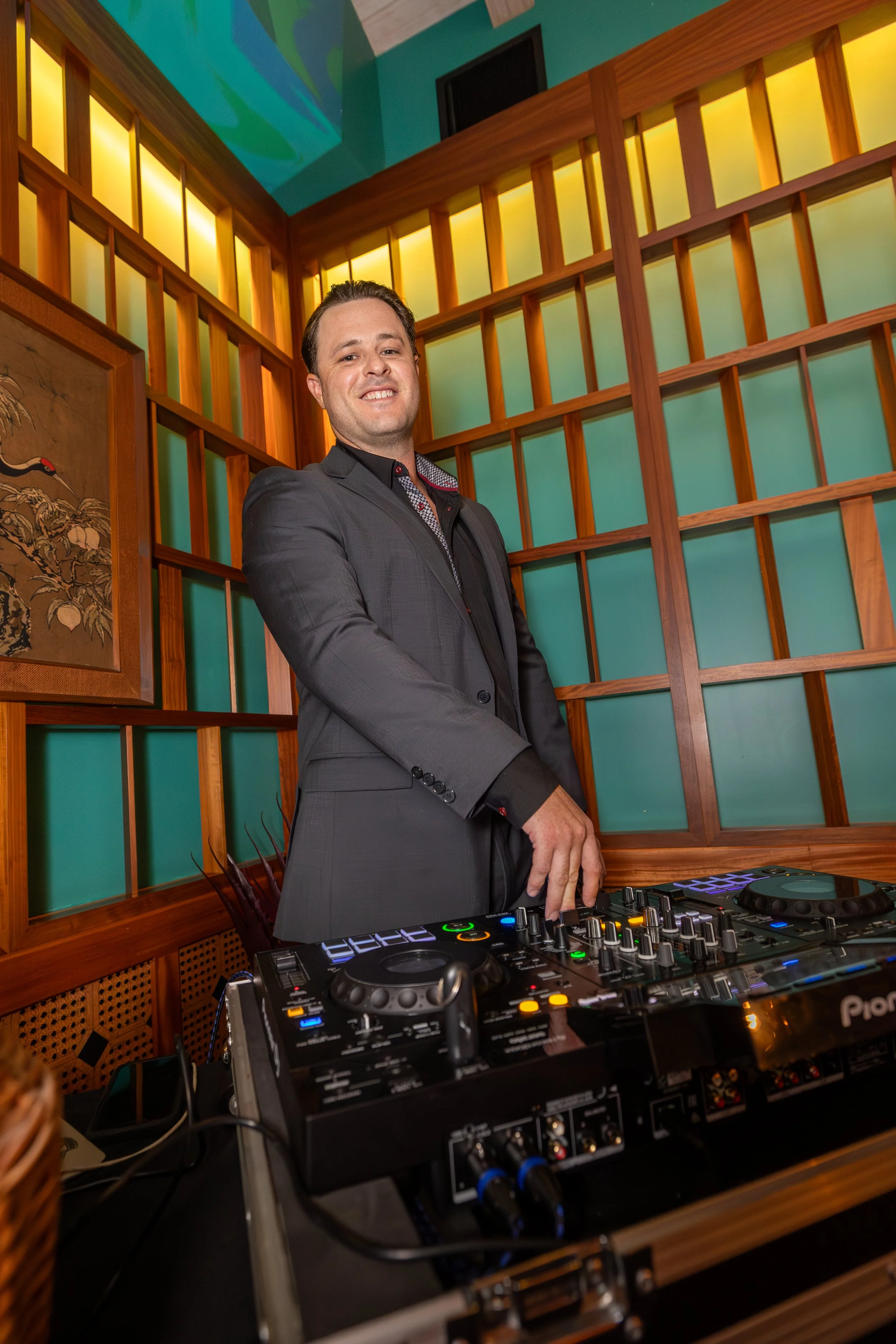 A man in a black suit standing behind DJ equipment in a wedding with wooden accents and colorful walls.