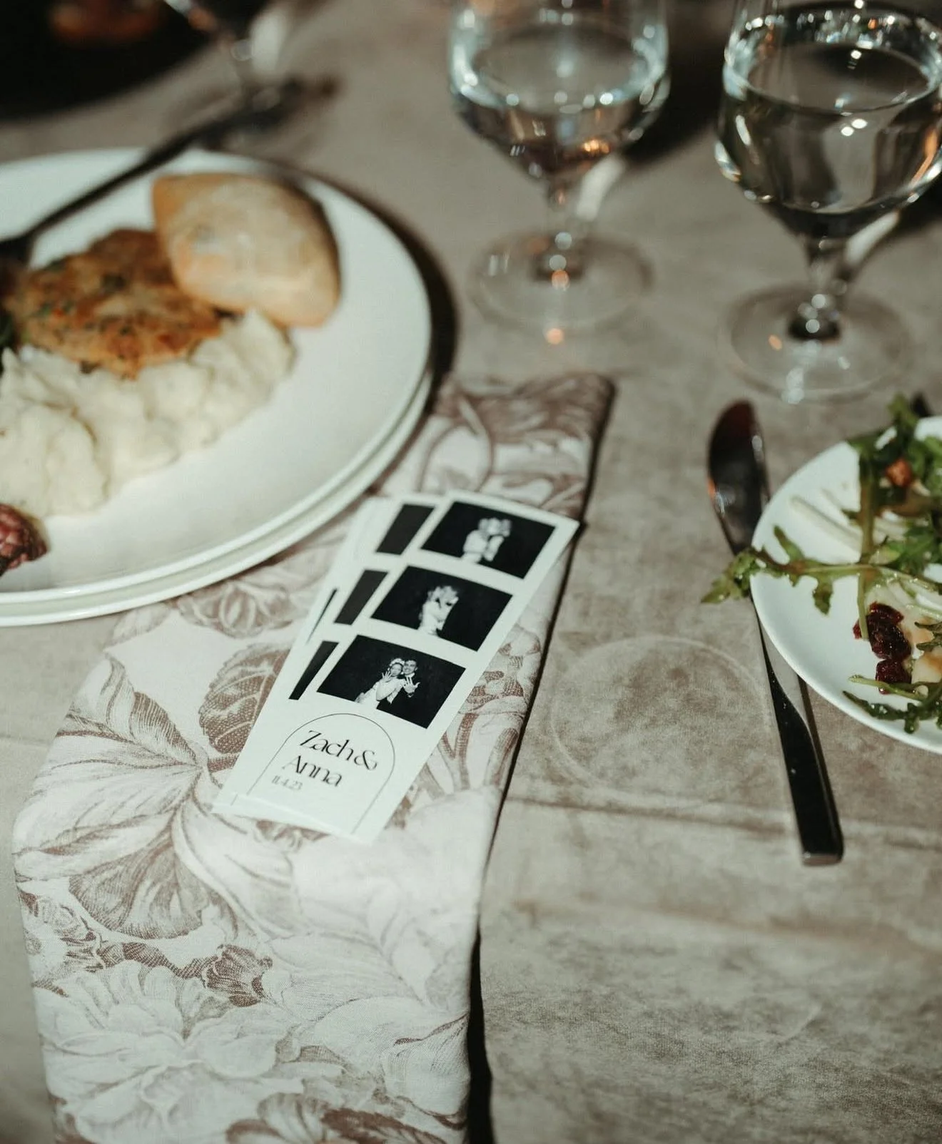 A dinner table setting with plates of food, two glasses of water, and a photo booth strip on a floral-printed napkin.