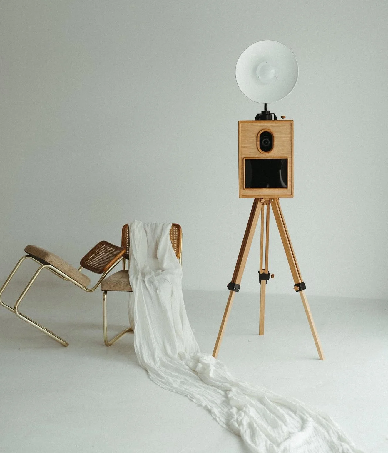 A vintage-inspired photo booth setup with a wooden tripod standing camera, a large white studio light, a beige chair with a cane backrest and metal legs, and a white fabric draped over the chair, all against a plain light gray background.
