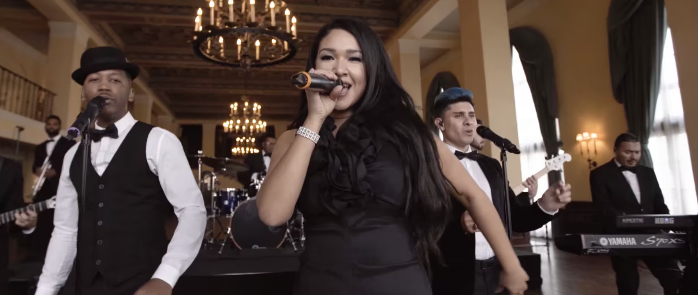 A live band performance in an elegant room with chandeliers and draped curtains, featuring a female singer in a black dress holding a microphone, surrounded by male band members in formal attire, some playing instruments.