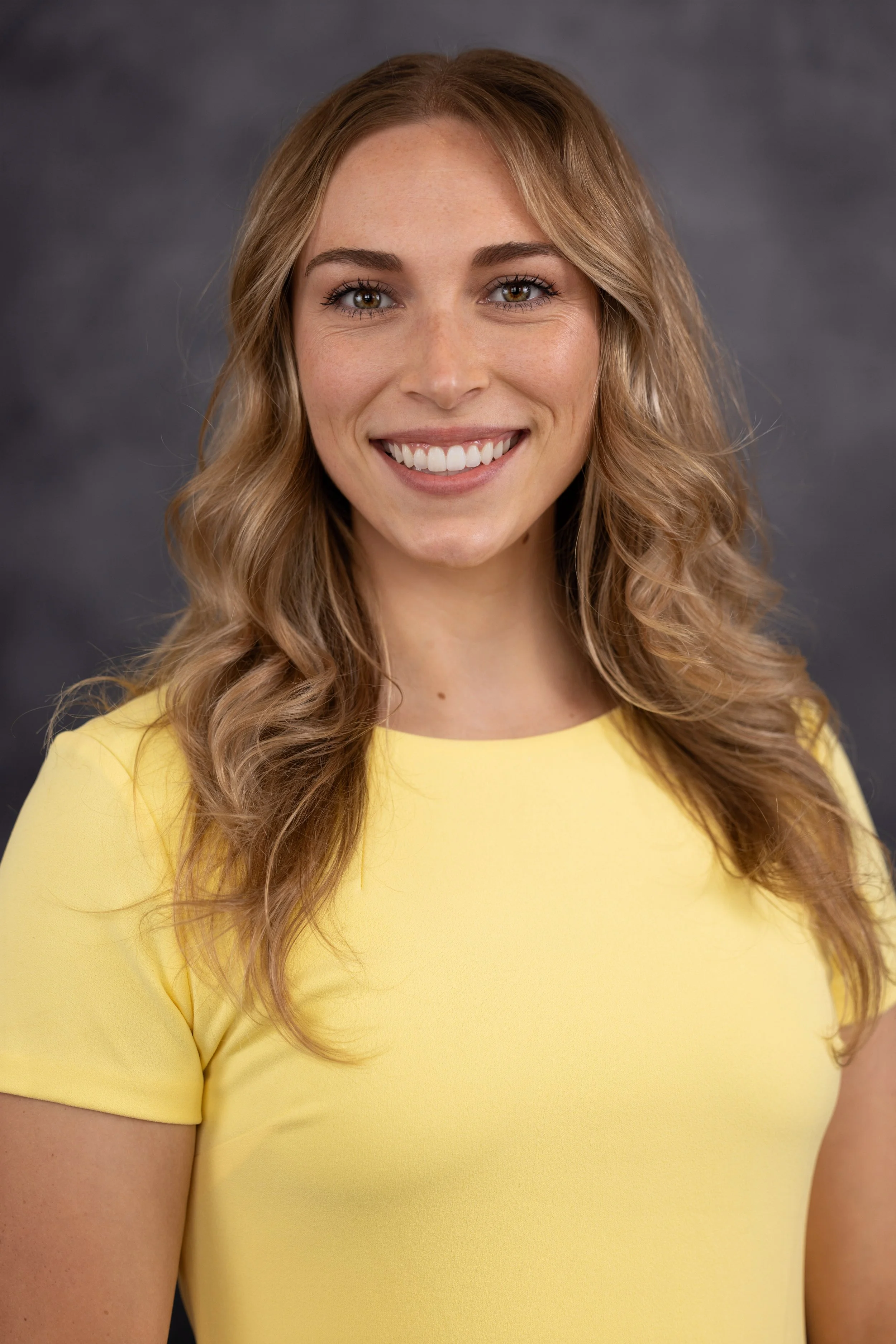 A woman with long wavy light brown hair and light green eyes, smiling with bright white teeth, wearing a yellow short-sleeved dress, posing against a gray background for a professional headshot.