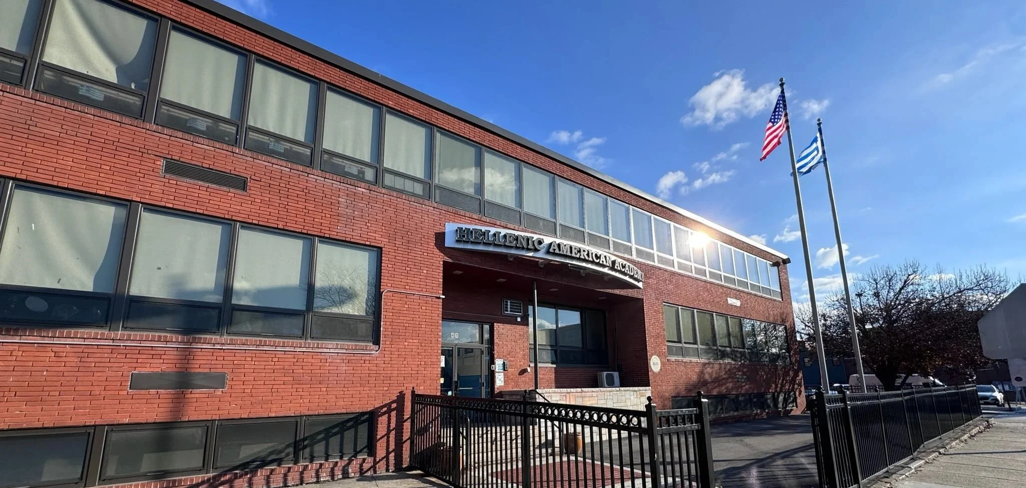 Red brick building with large windows and a sign that reads 'Hellenic American Academy'. Two flags, the American flag and the Greek flag, are flying on flagpoles in front of the building. The sky is blue with some clouds.