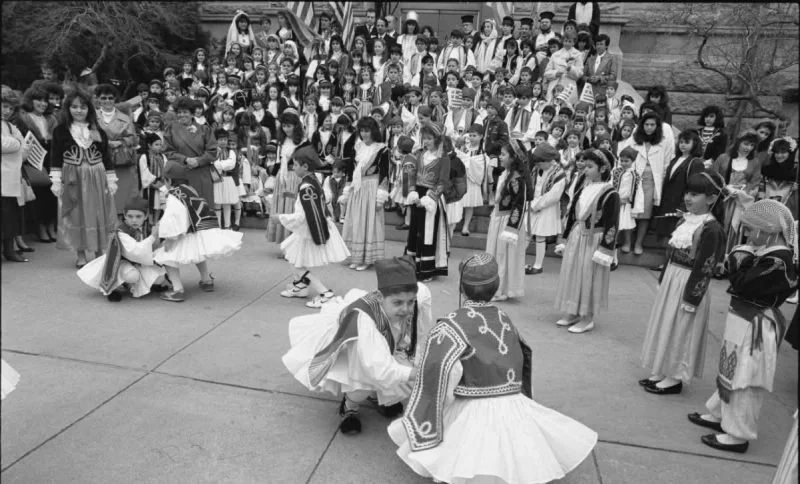 Children in traditional costumes participating in a cultural dance outdoors, with a large group watching from stairs and the background.