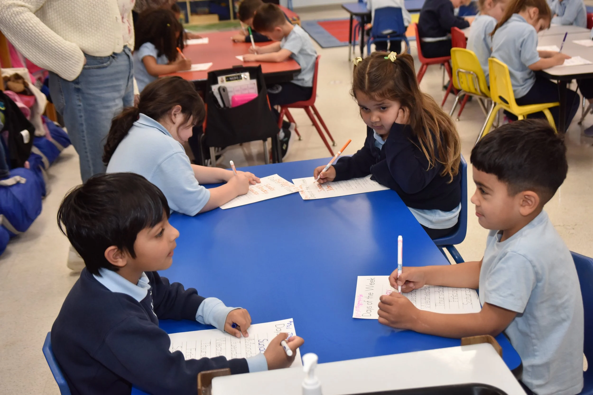 A classroom filled with young students sitting at tables, working on assignments with pens and paper.