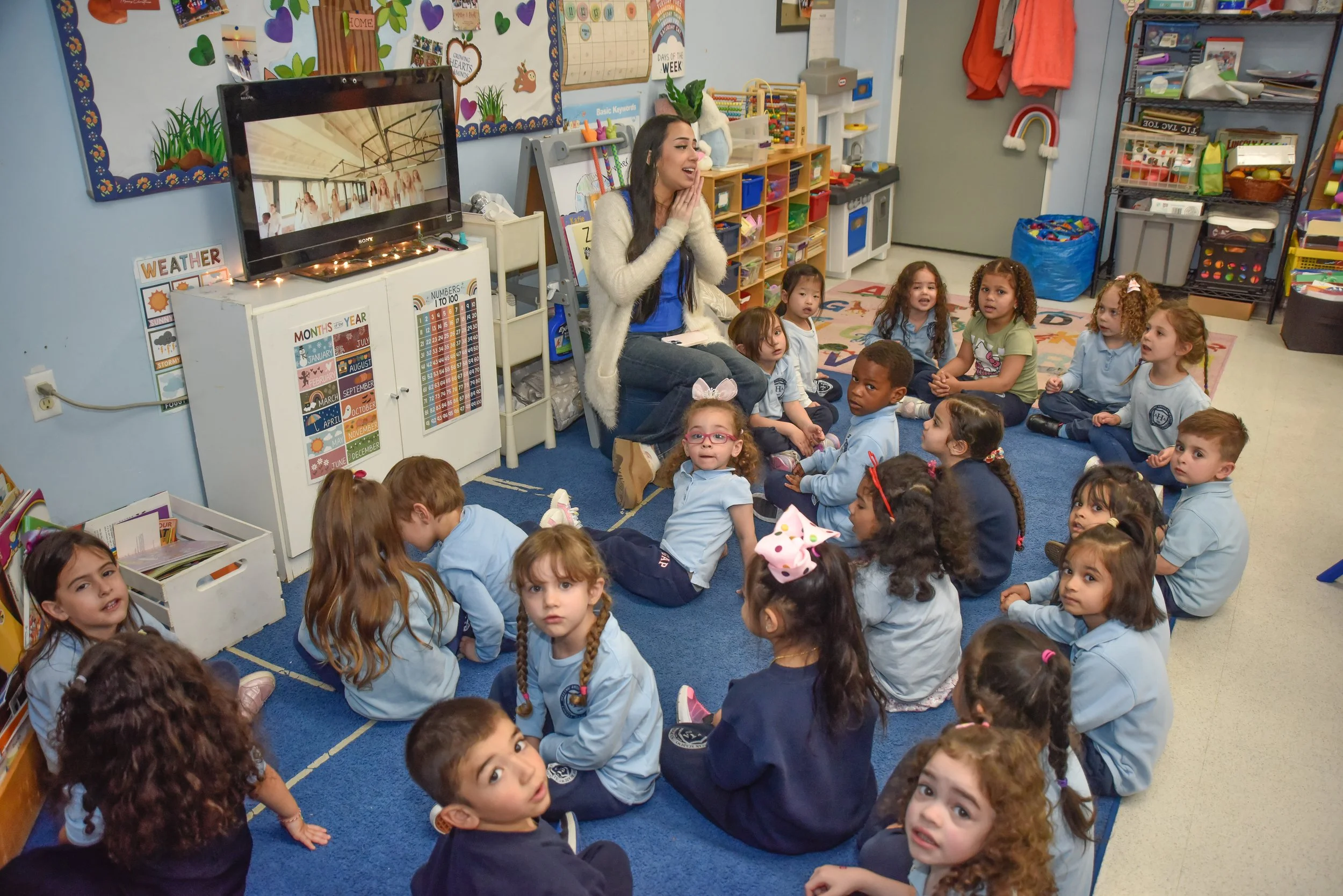 A teacher sitting on a small chair in front of seated children in a classroom. The children are sitting on a blue carpet, and the classroom has shelves with educational materials, a television, and colorful decorations on the walls. The teacher is smiling and talking to the children.