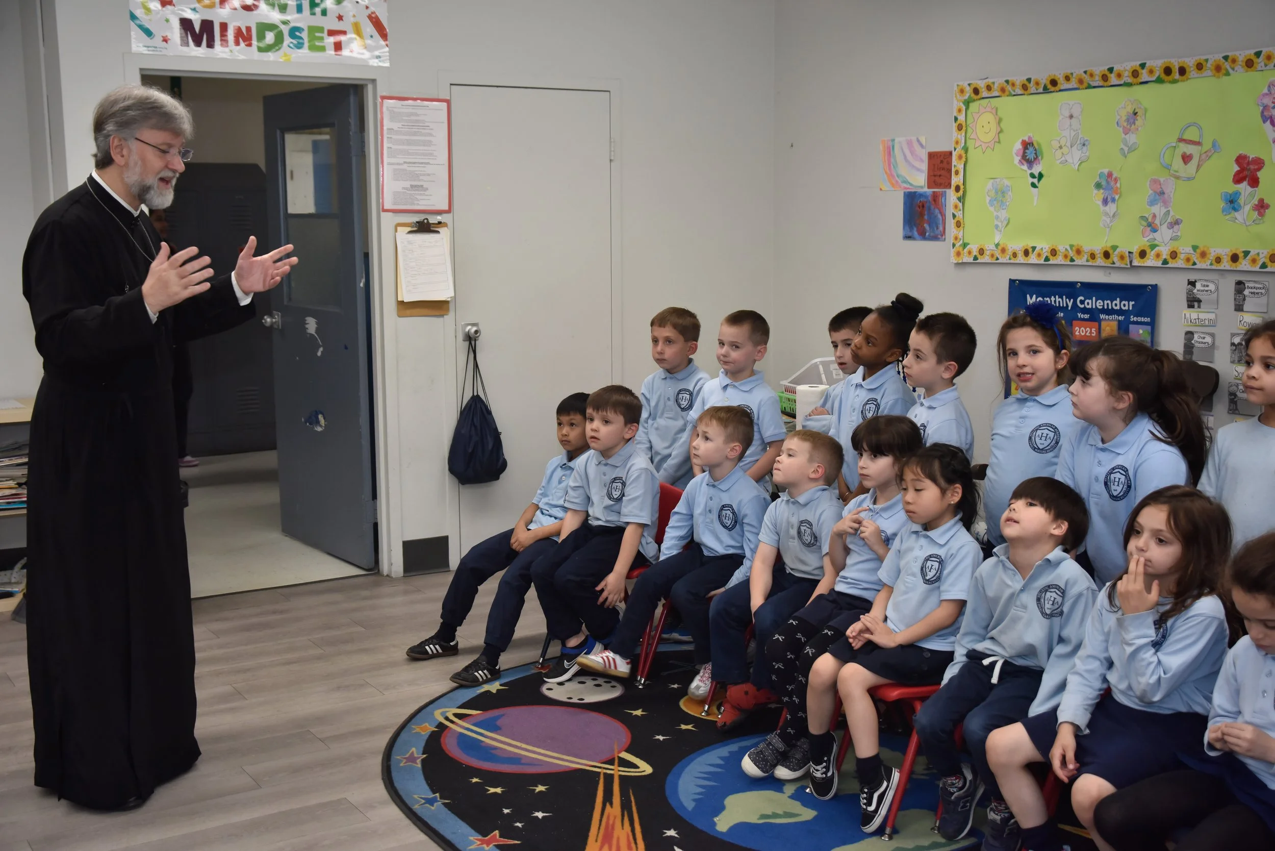 A man with gray hair and a beard, dressed in black robes, is talking to a group of children seated on chairs in a classroom. The children are wearing light blue uniforms with a school logo and are attentively listening. The classroom has colorful decorations, a school calendar on the wall, and a space-themed rug on the floor.