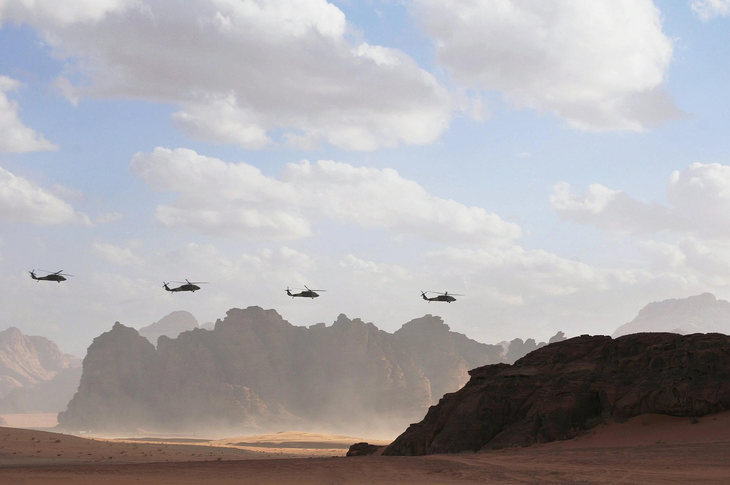 Four military helicopters flying over a desert landscape with rocky formations and cloudy sky.