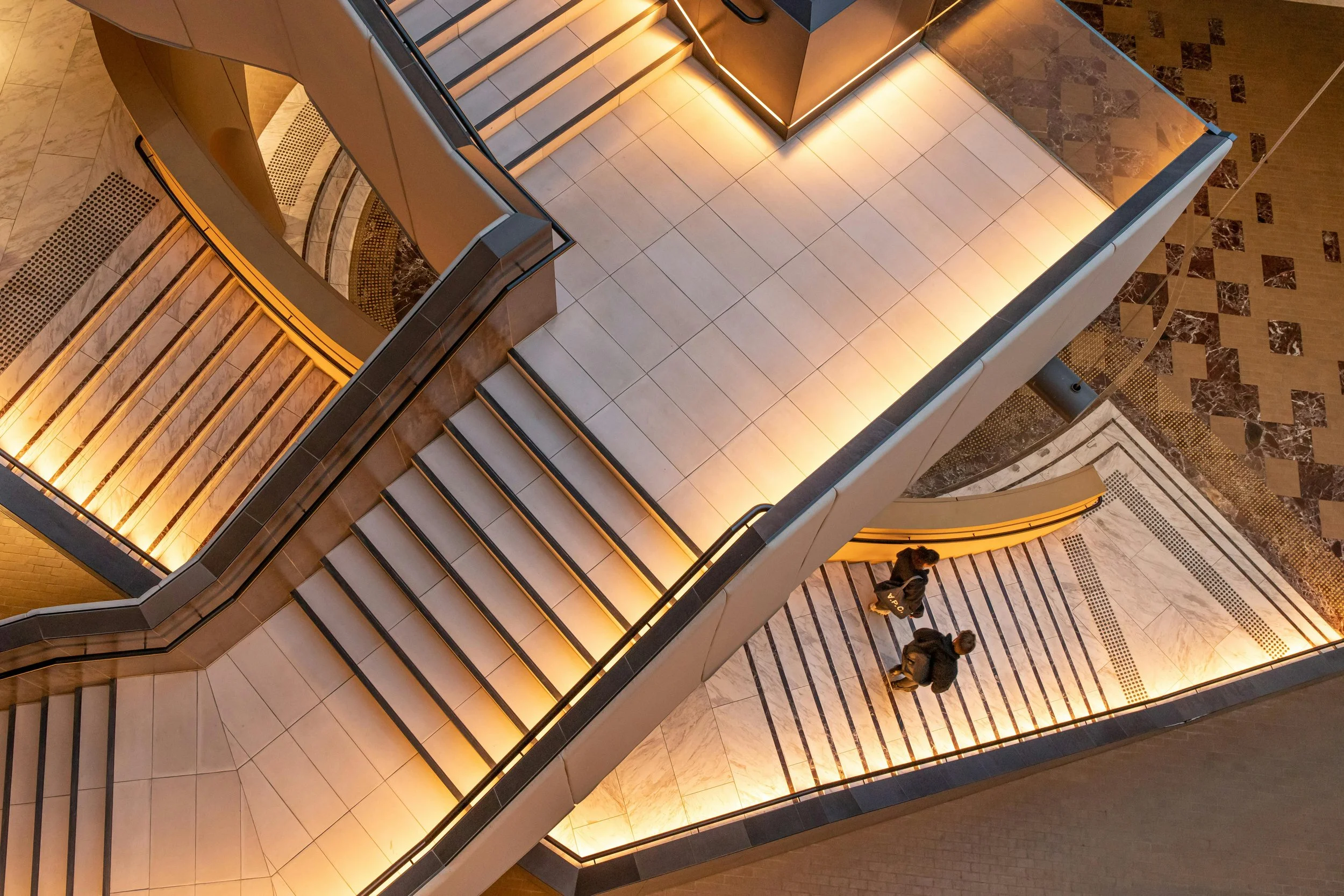 Two people walking down a staircase in a modern building, viewed from above, with warm lighting and tiled floors.
