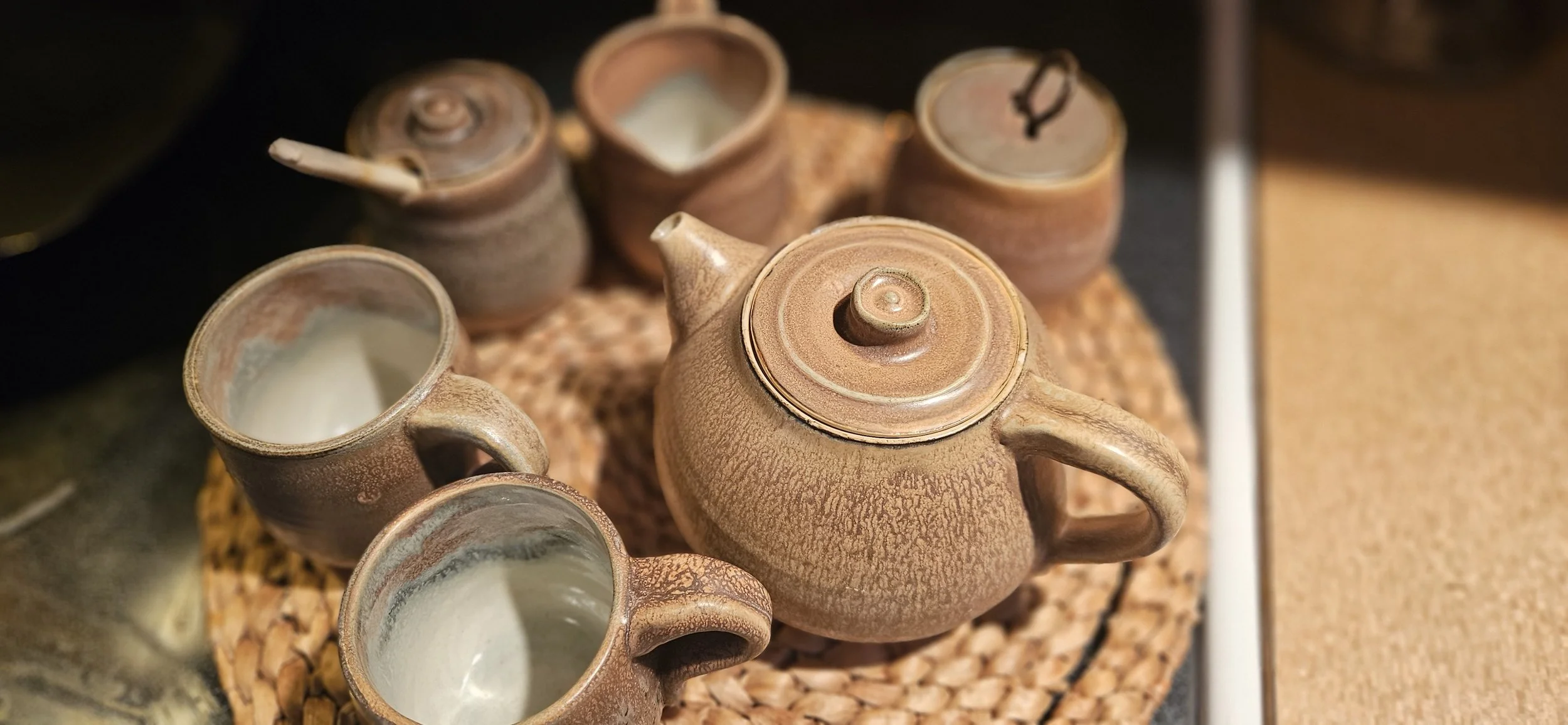 A ceramic teapot and several matching cups with white liquid, arranged on a woven mat.