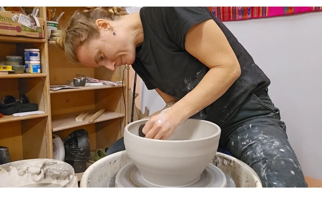 A woman shaping a ceramic bowl on a pottery wheel in a studio with shelves of supplies.