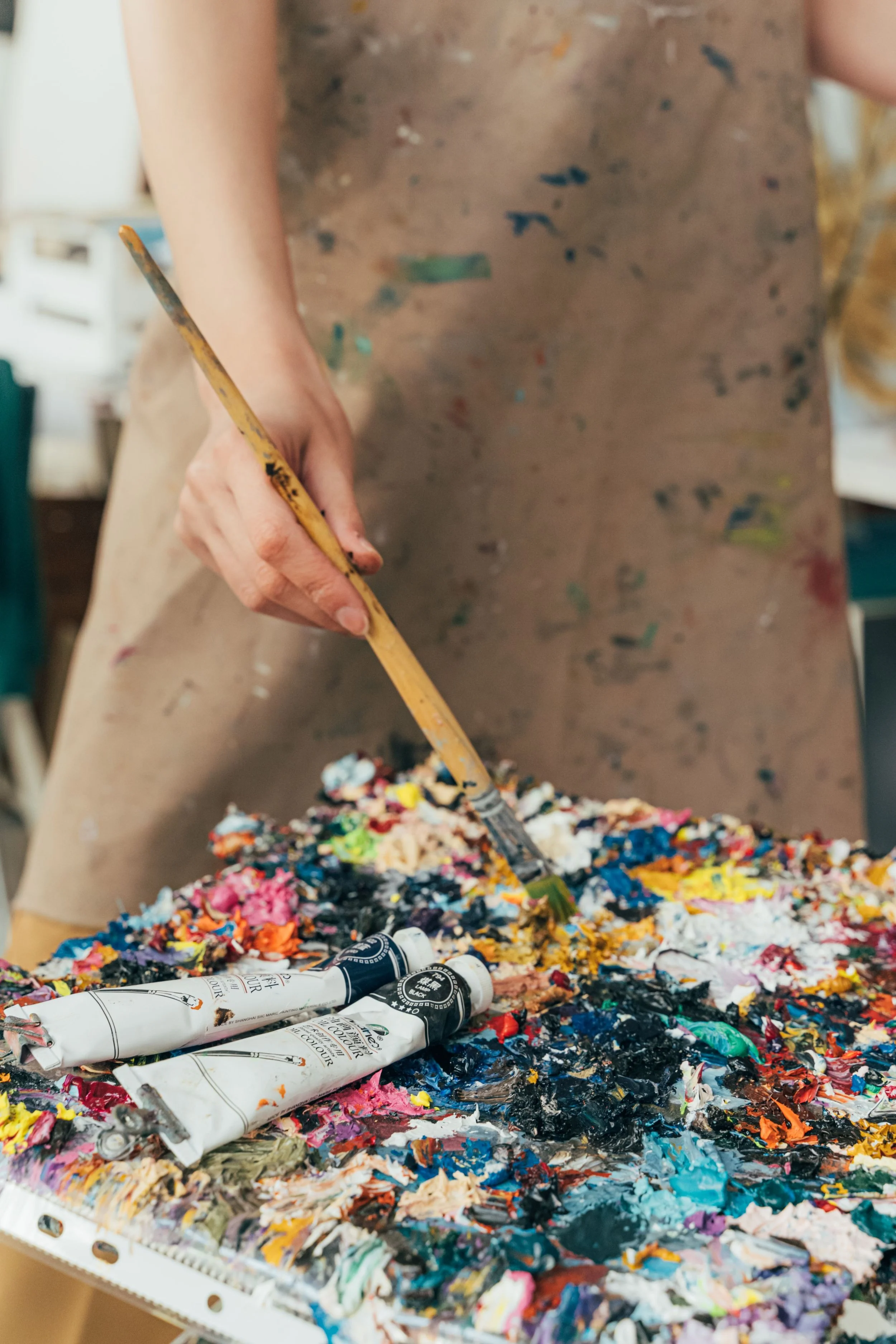 An artist's hand holding a paintbrush over a messy palette of oil paints in various colors, with paint tubes nearby.