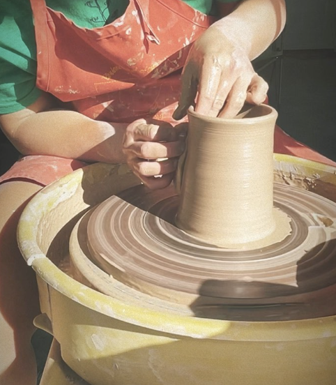 A person shaping clay on a pottery wheel with their hands, wearing a red apron and green shirt.