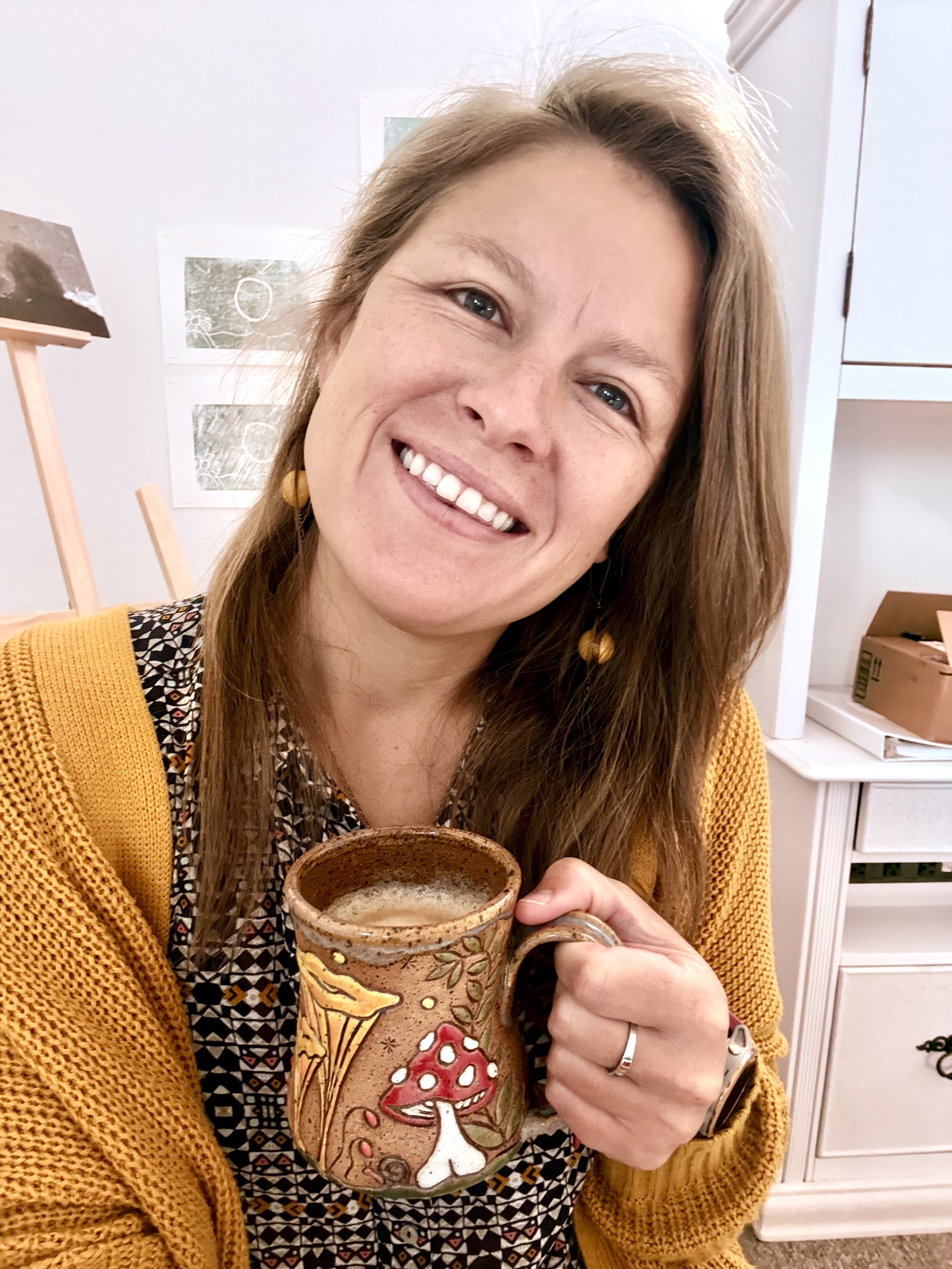 A woman with long hair, wearing a yellow cardigan and patterned dress, holding a decorated ceramic mug with a mushroom drawing, smiling in a kitchen.