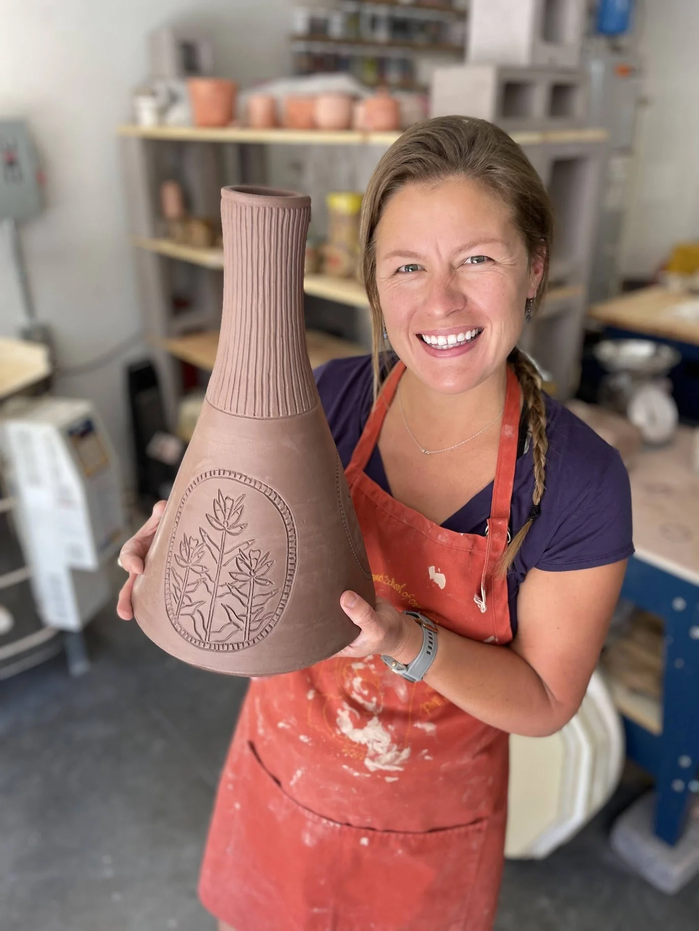A woman with long hair tied in a braid, smiling, holding a large, unglazed ceramic vase with a textured, ridged top and engraved leaf pattern on the body, in a pottery studio.
