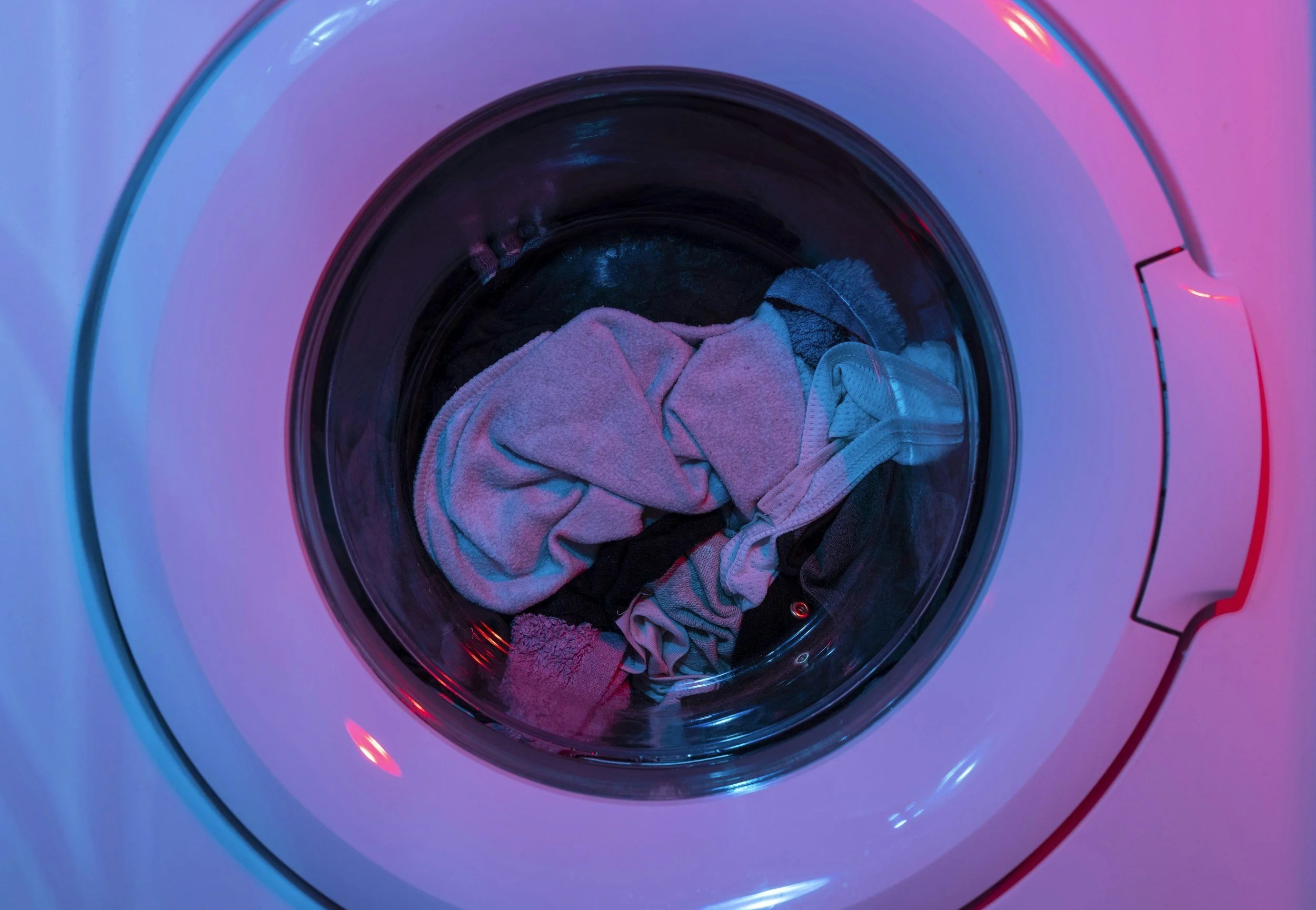 A front-loading washing machine with clothes inside, illuminated by pink and blue LED lights.