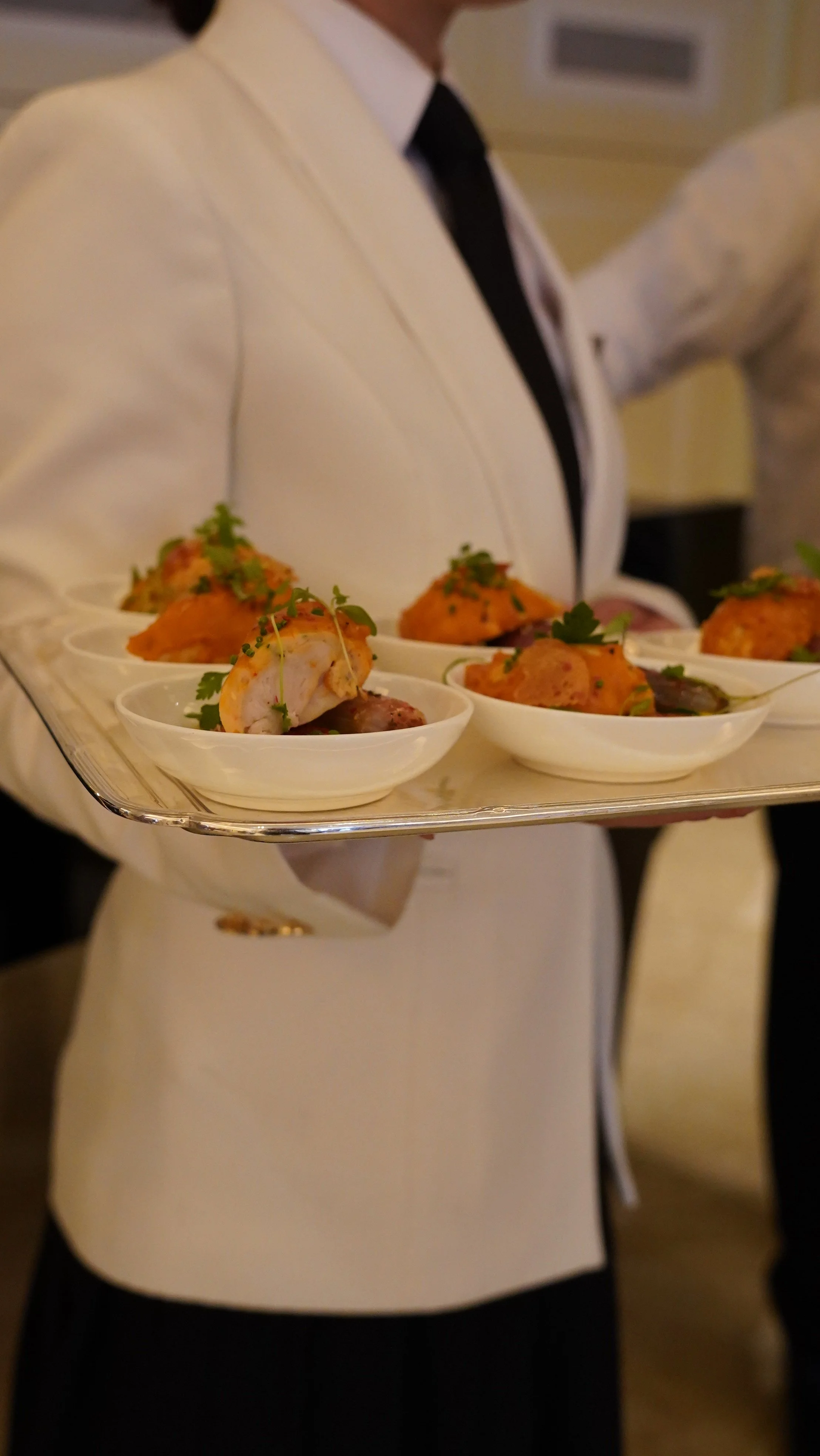 A person dressed in a white suit jacket with a black tie holding a tray of appetizers with small bowls of food garnished with herbs.