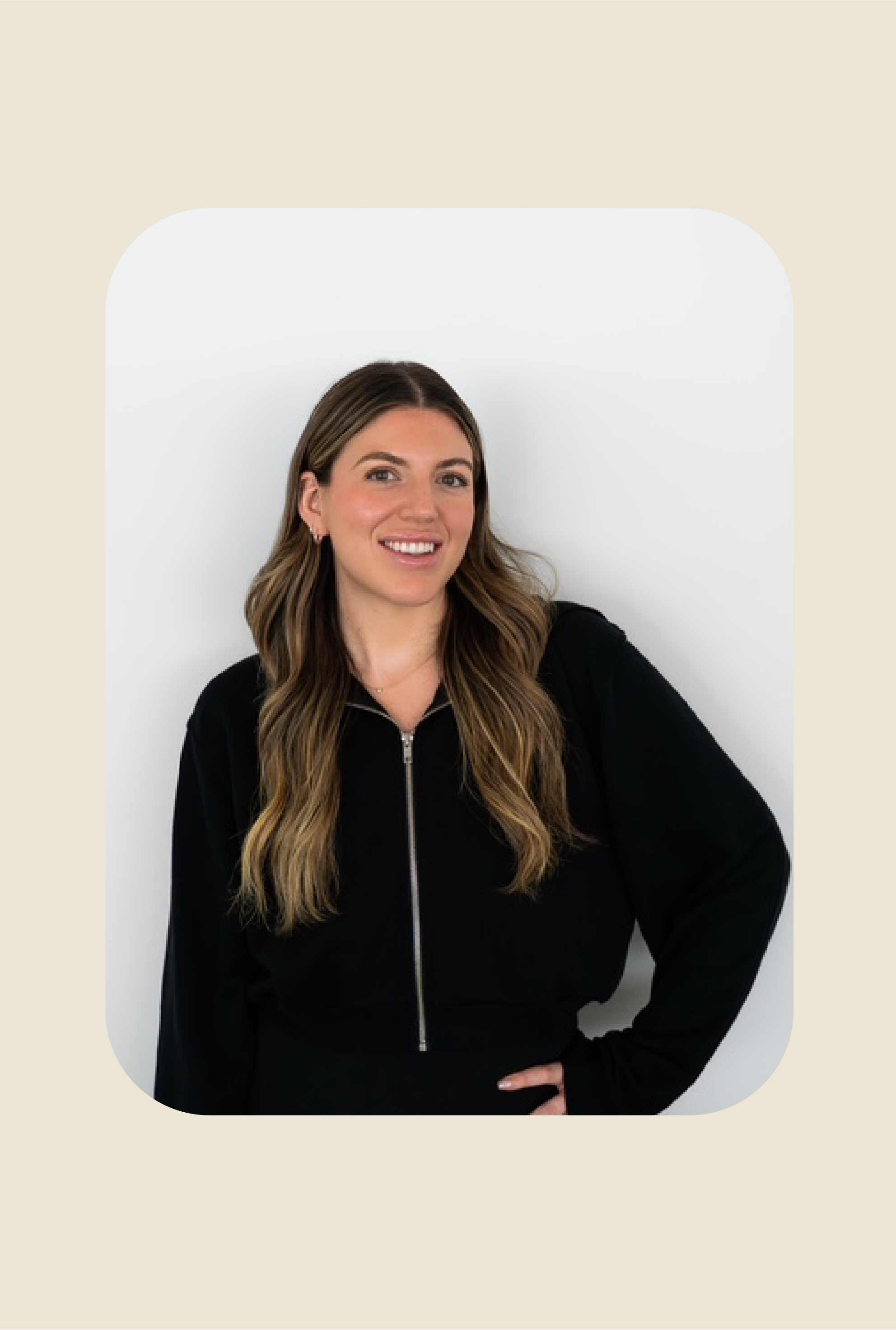 A woman with long, wavy brown hair, wearing a black zip-up jacket, smiling and posing with her left hand on her hip, standing against a white background.