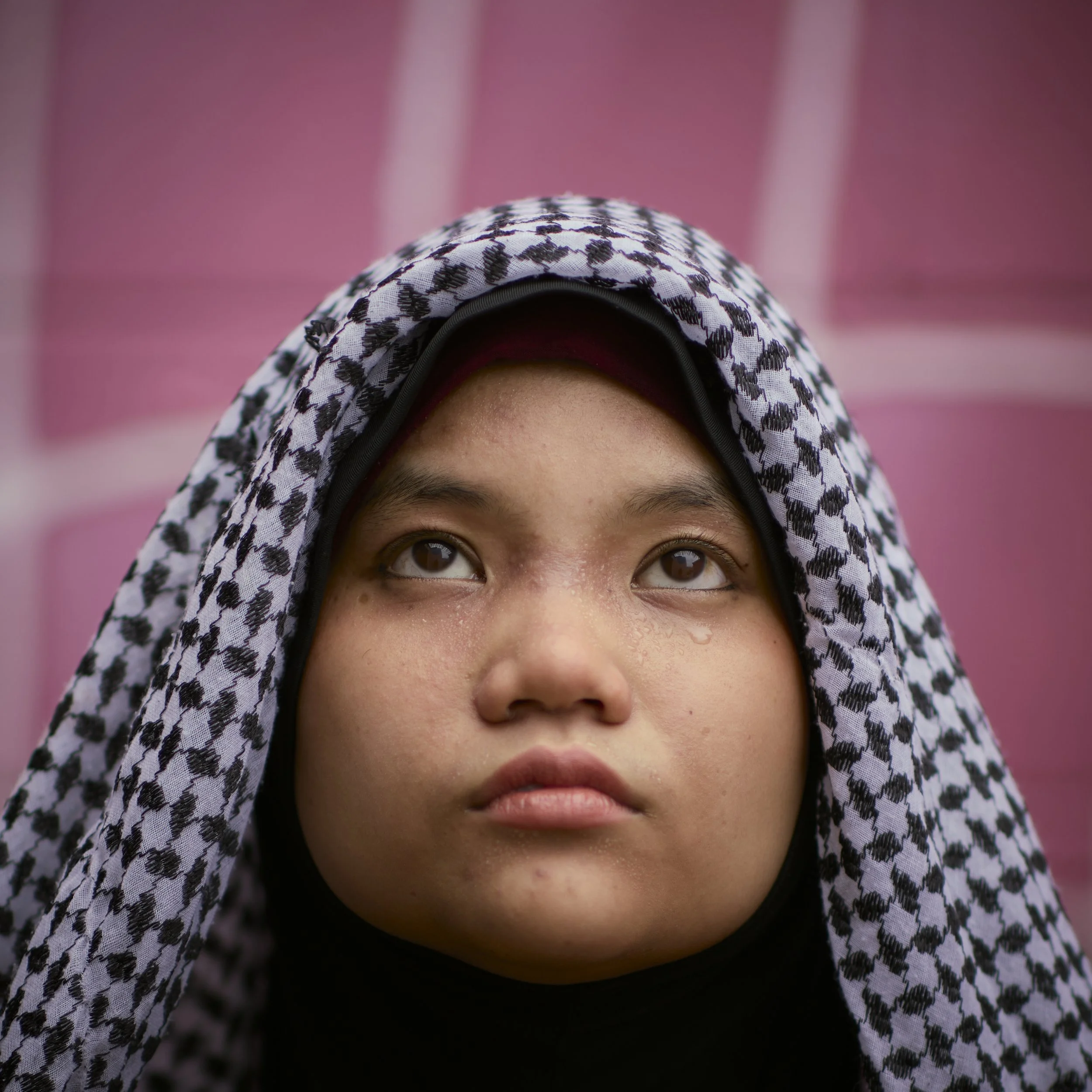 Close-up of a young person wearing a black and white keffiyeh scarf, looking upwards with a somber expression, against a pink background.