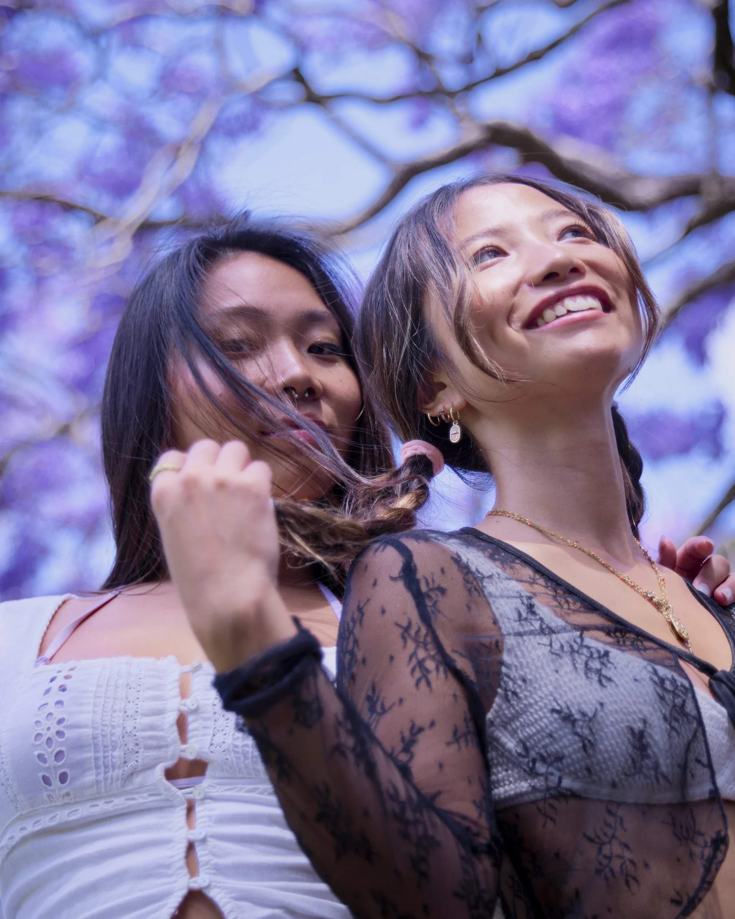 Two young women smiling outdoors, with purple flowering tree branches and blue sky in the background.