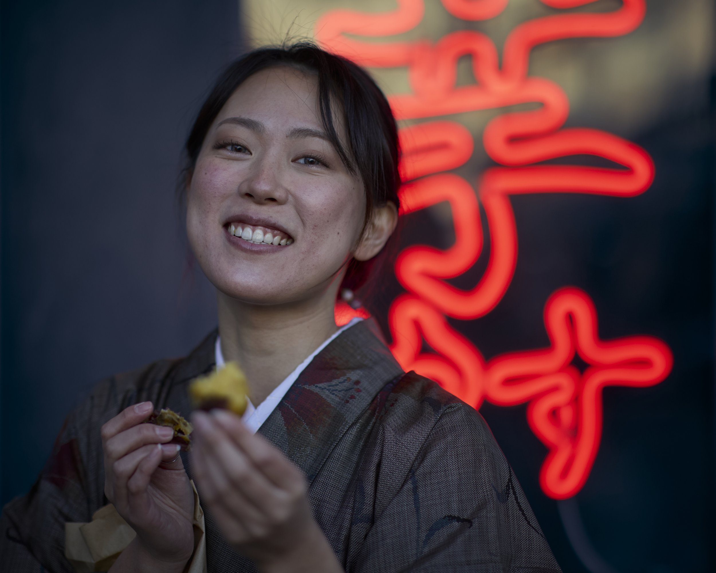 A young Asian woman smiling and holding a piece of food in her hand, standing in front of a dark background with an illuminated red neon sign.