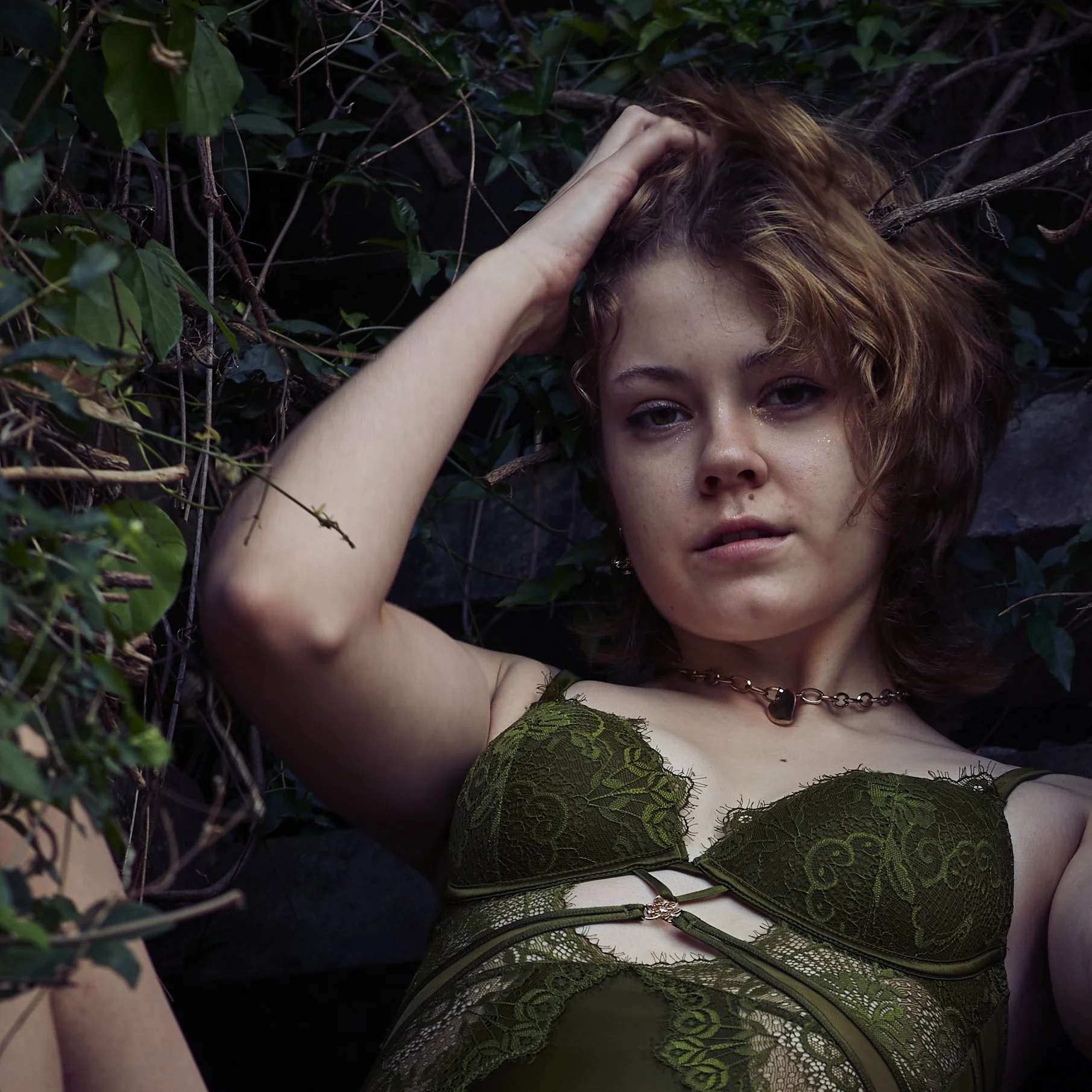 A young woman with wavy, reddish-brown hair lying among dark green ivy leaves, wearing a green lace dress and a choker necklace, looking directly at the camera.