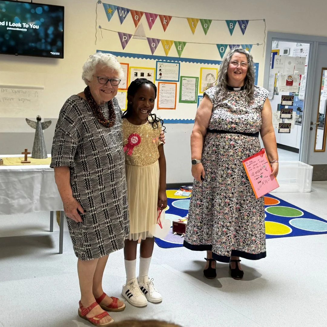 Three women and a girl standing indoors, smiling for a photo. Behind them, colorful decorations and a notice board are visible, indicating a school or community event. The girl is wearing a dress with a badge, and the women are dressed in patterned dresses.