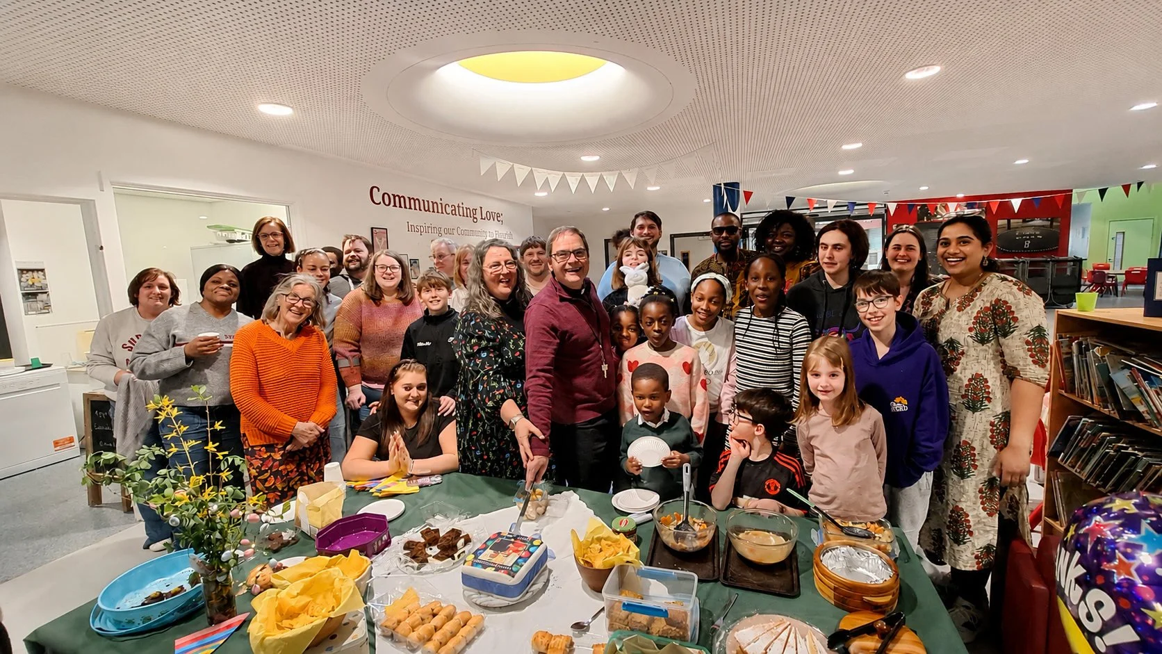 Group of diverse people, including children and adults, gathered around a table with food in a community room, celebrating together.